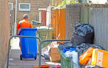 Refuse worker pulling wheelie-bin along an alley filled with rubbish.