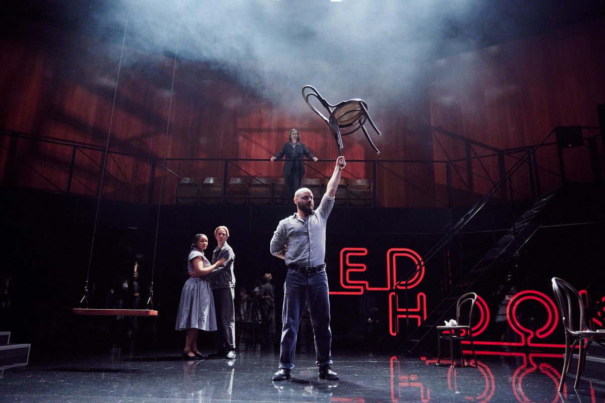 A dimly-lit theatre stage with a bald, white man front and centre, holding a wooden chair up over his hand with his left hand. Behind him to his left: a woman and a man holding each other. Behind him to his right: red neon lettering. Further back up on a gallery is a person holding the railing and looking down at the scene