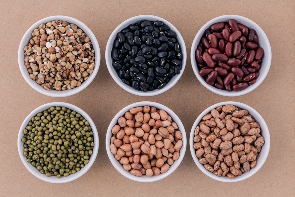 Bird-eye view of six white bowls filled with different kinds of pulses on a tan surface. 