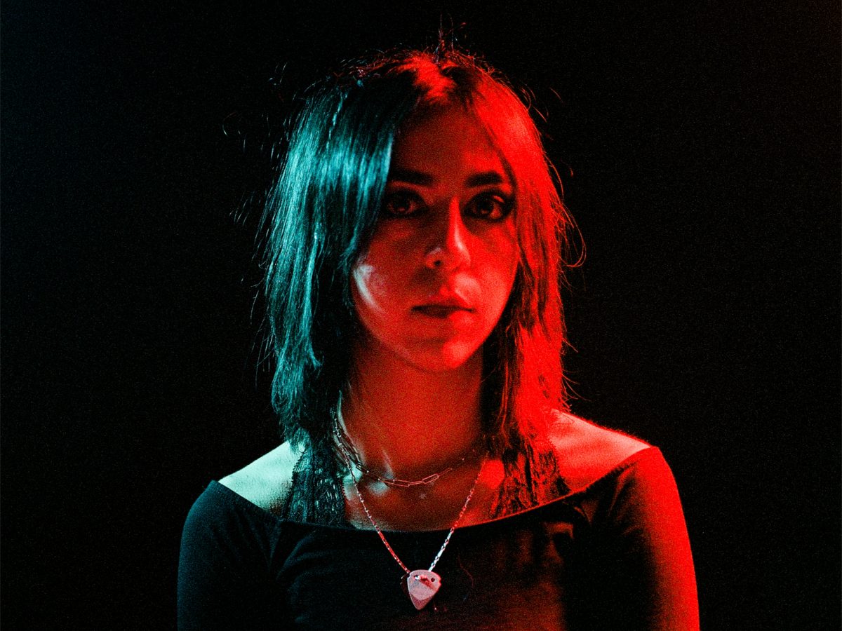 Portrait of Nadia Sheik, a you British-Spanish indie artist. The white woman has wavy brown hair and is looking at the camera. The right side of her face is illuminated in red light.