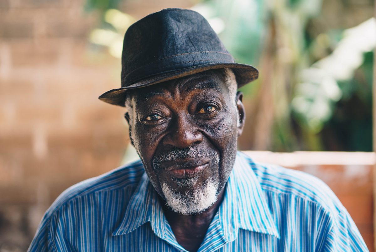 A portrait of an older black man with a grey beard. He’s wearing a brown hat and a white and blue striped shirt. He’s looking at the camera with a friendly, neutral expression. The background is blurry.