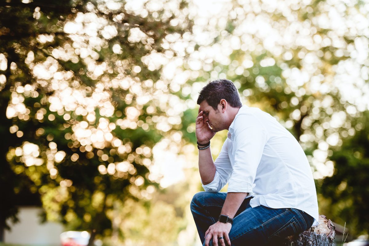 A white man in a white shirt and blue jeans is sitting in nature, holding his head in his hand, looking sad and distressed. The threes in the background are blurred out.