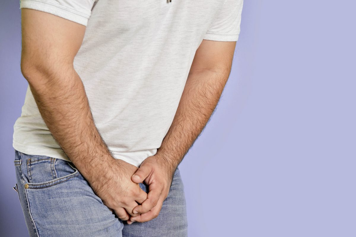 A close-up of a white man holding his crotch. He’s wearing a white t-shirt and jeans, the background is lilac. The shot shows him from under his shoulders to the top of his thighs.