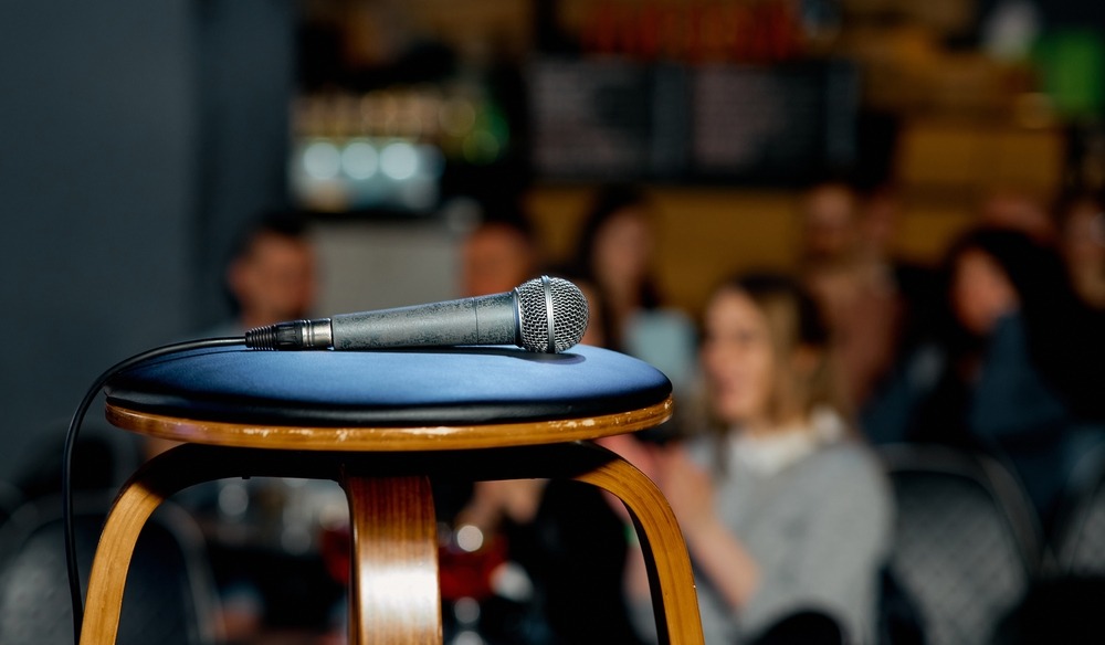 A stool with a microphone in the foreground. Behind it you can see a small audience, clapping and laughing, but blurred. 