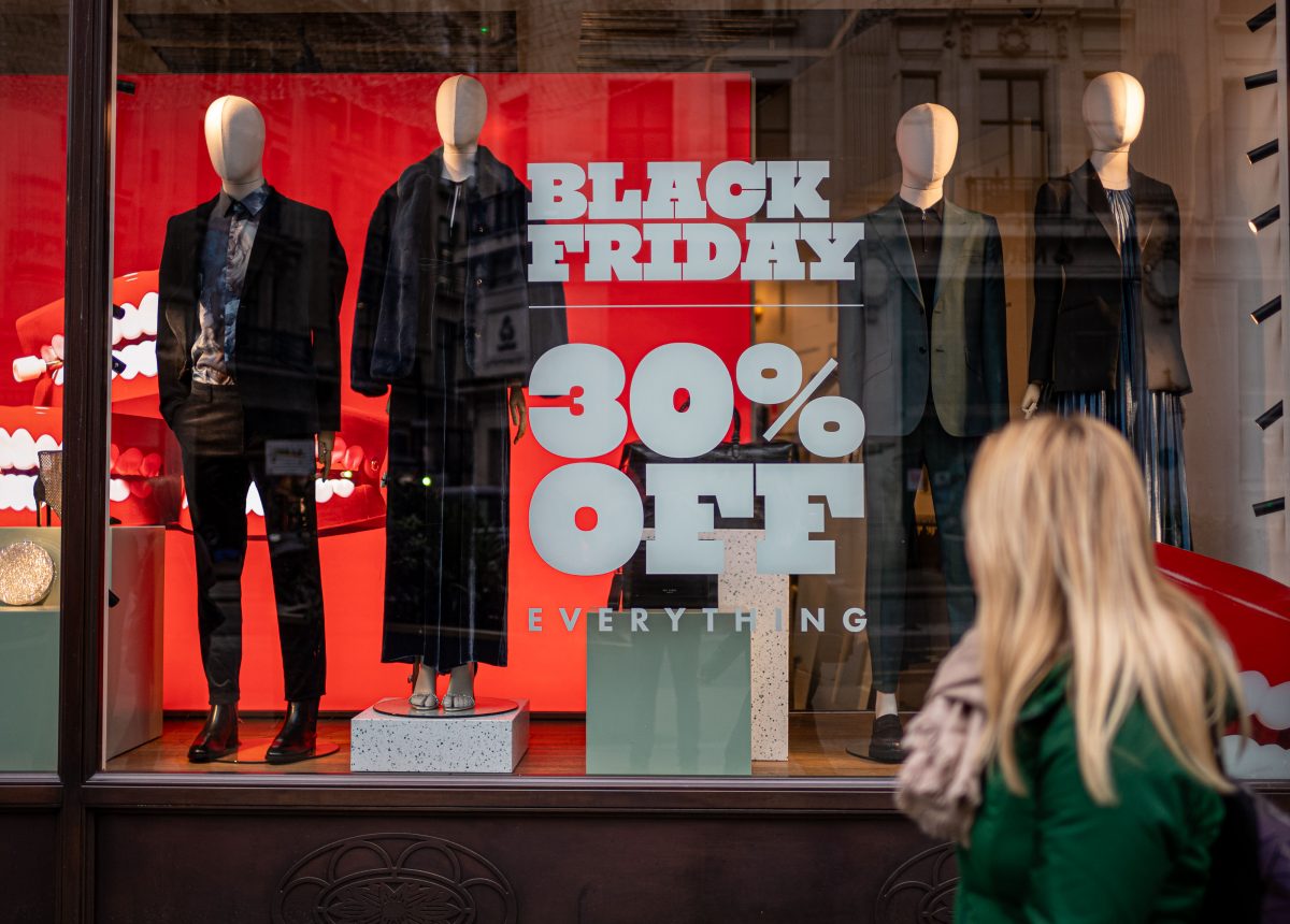 A woman with long blonde hair in front of a shop window. She has her back to us and is looking at the shop behind her. Inside are mannequins and grey letters on the window read: “Black Friday, 30% off everything”