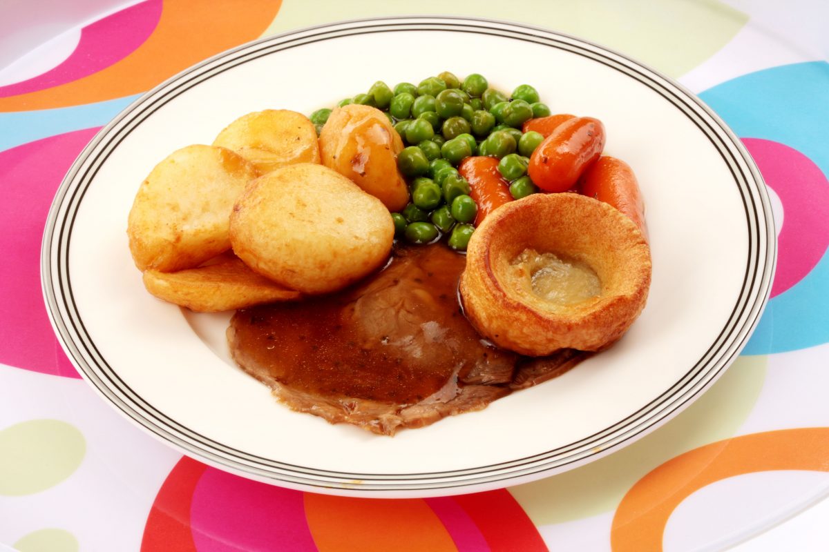 A plate of roast lamb with a Yorkshire pudding, roast potatoes, peas, carrots, and gravey