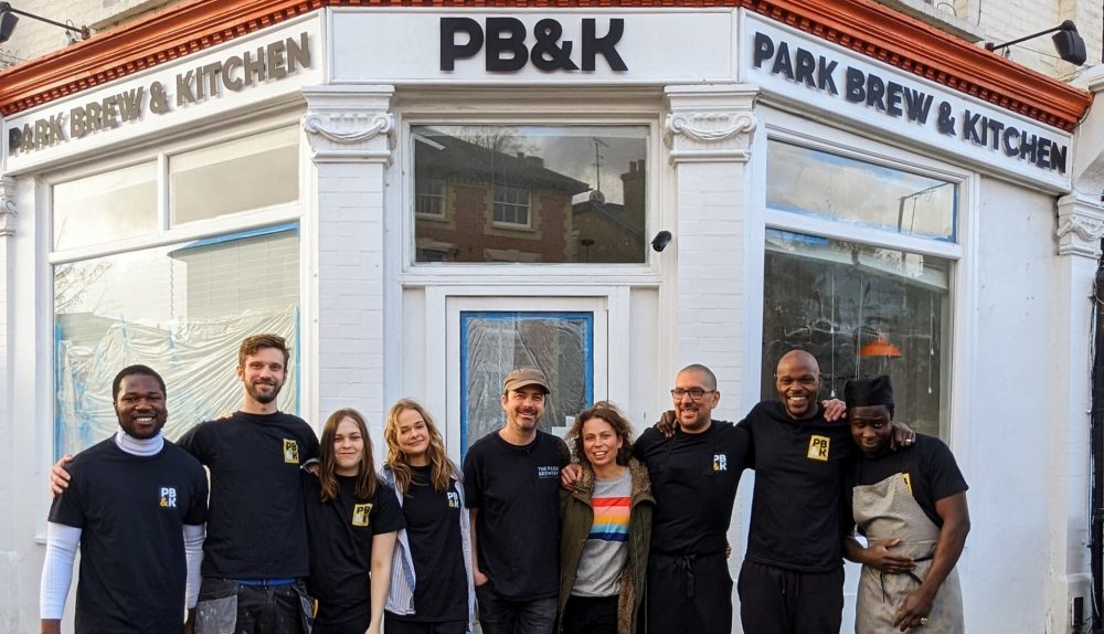 A team of nine people in black t-shirts standing outside a white corner shop with the following lettering: PB&K