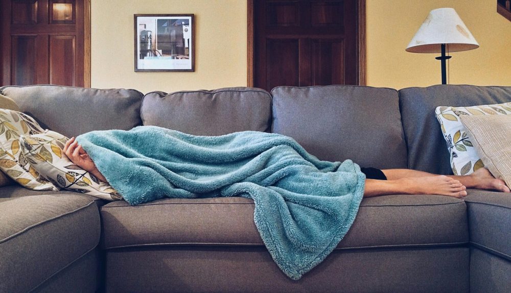 Sick person laying on sofa with blanket over their body.