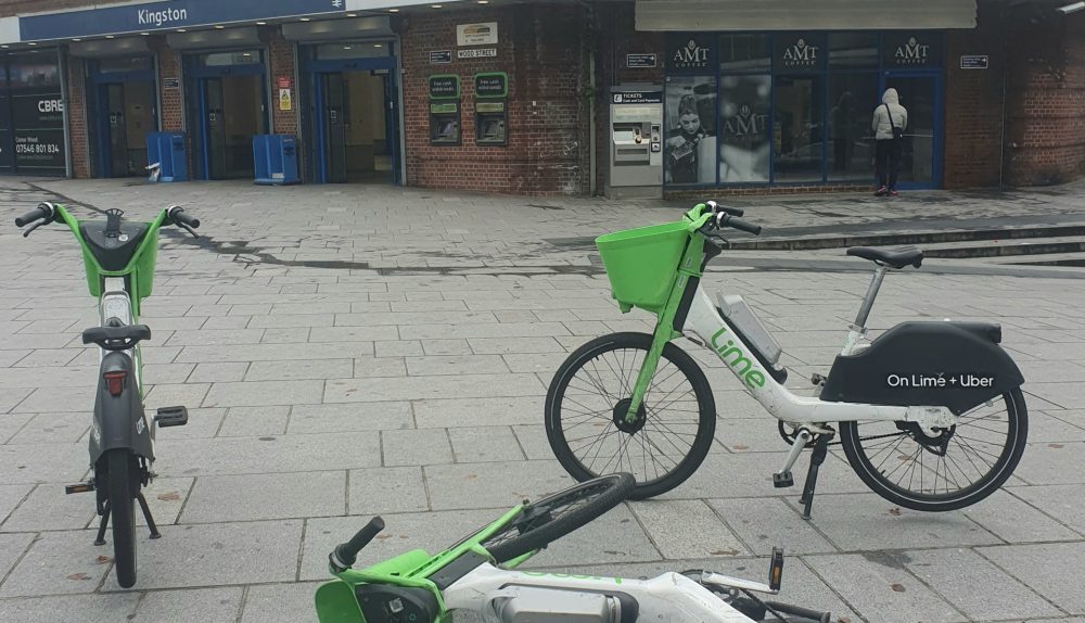 Three Lime bikes parked in the middle of the pavement in front of Kingston Station. One bike has fallen on the floor.