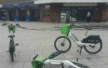 Three Lime bikes parked in the middle of the pavement in front of Kingston Station. One bike has fallen on the floor.