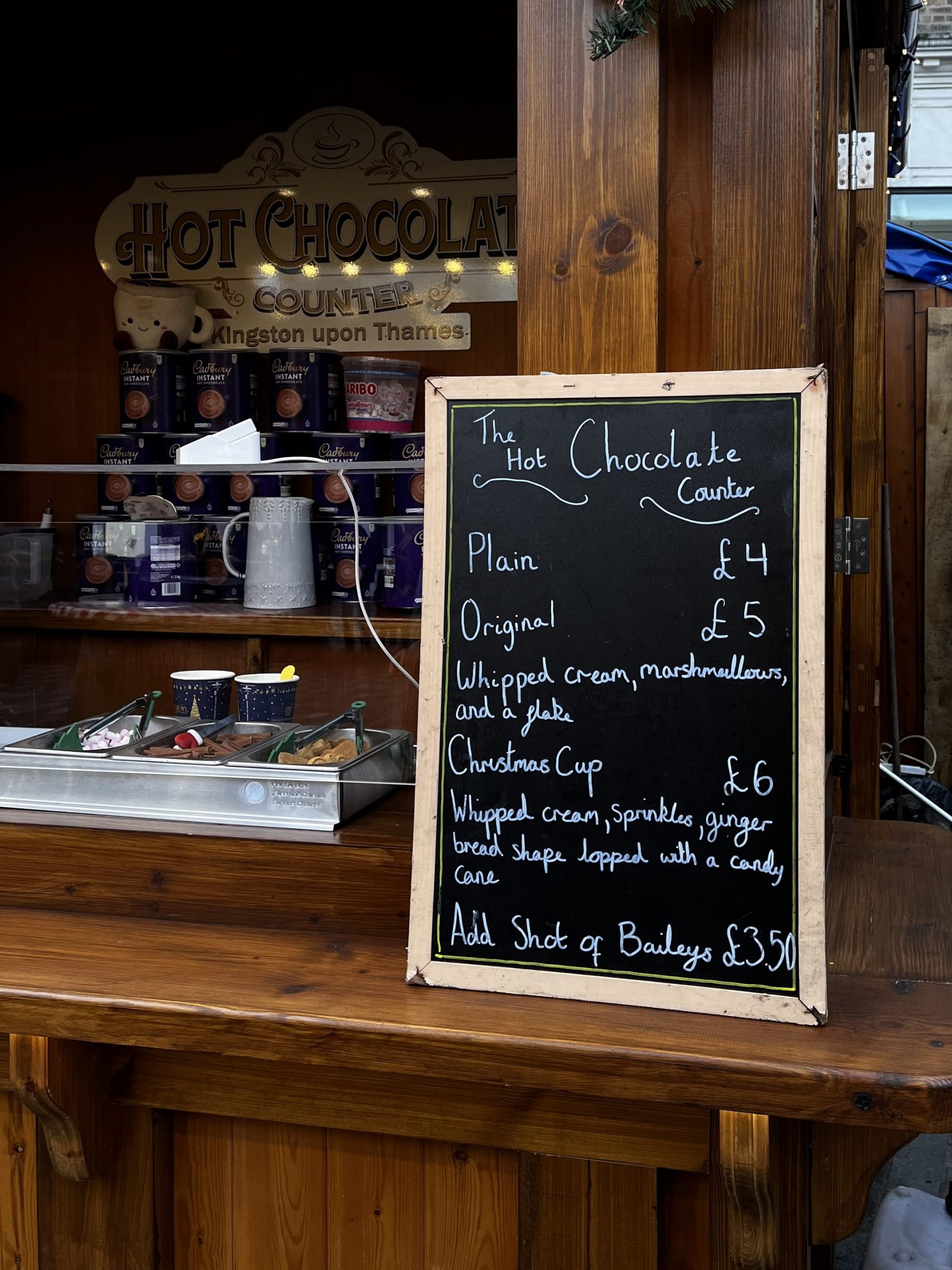 a menu of hot chocolate topping options, with a serving station of toppings and tongs in the backgroun